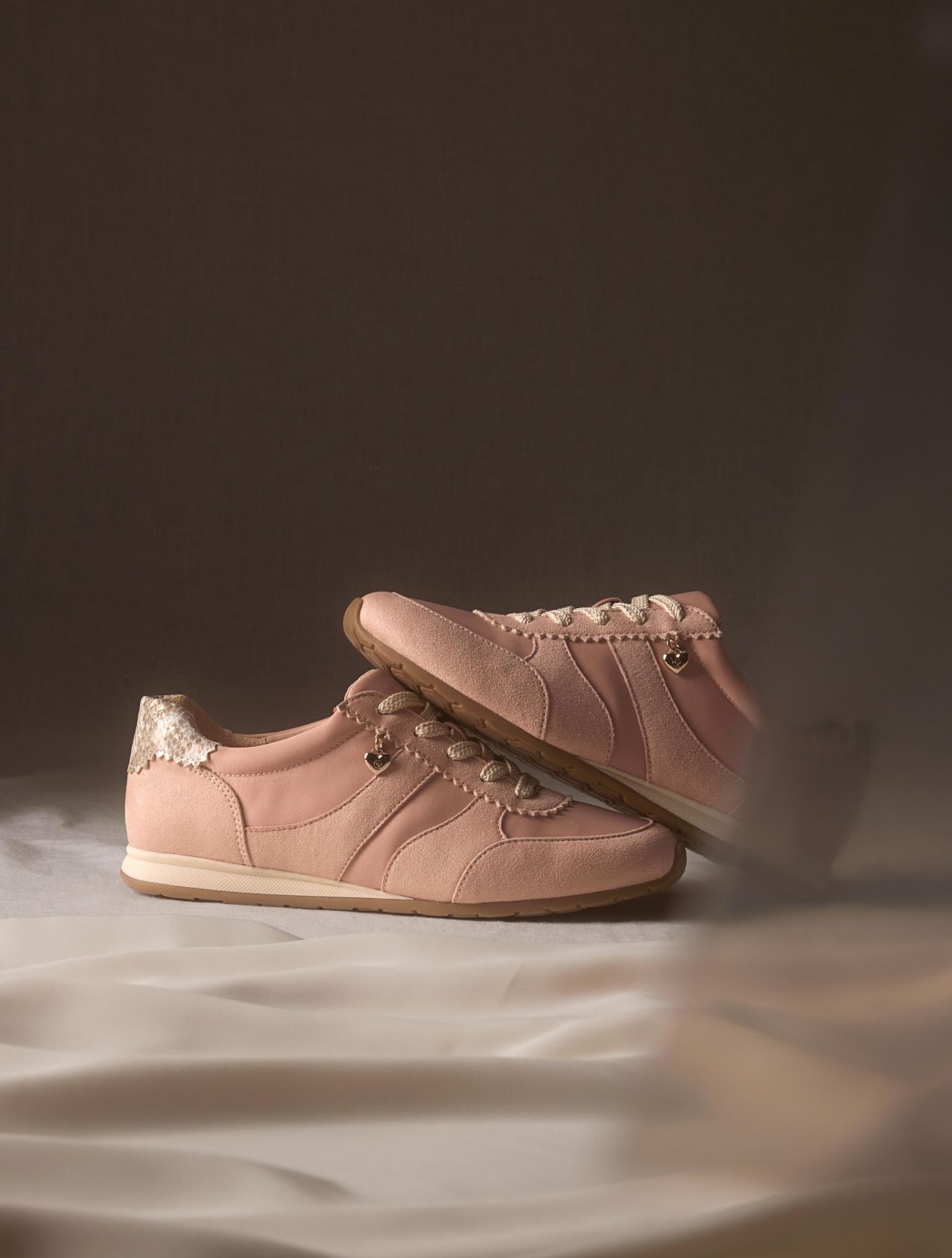A pair of blush pink sneakers with suede panels and metallic heel detail, set on a brown backdrop 