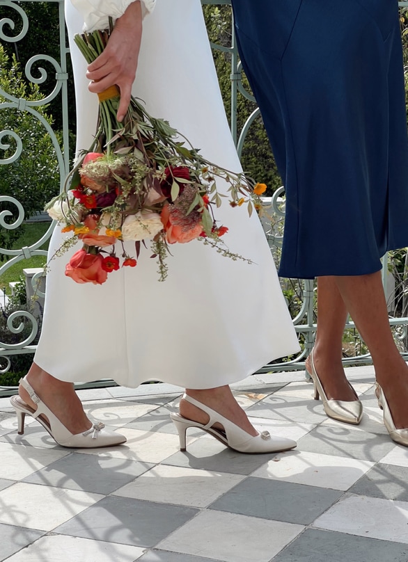 Two models in bridal outfits pose with a floral bouquet outside, wearing the gold metallic LifeStride Social pump and white satin Social Scene pump.