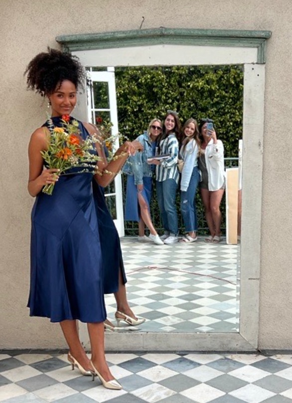 A model in a blue satin dress and the LifeStride Social pump holds a floral bouquet in front of a mirror reflecting a group of crew members.