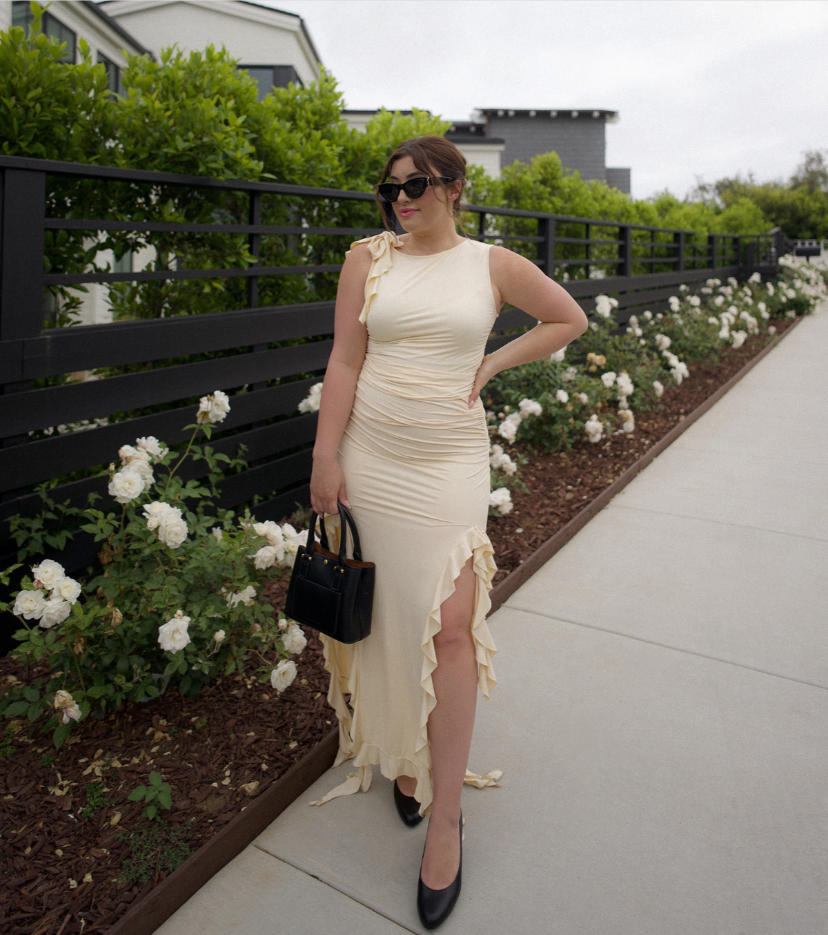 Influencer @katie_lavieri styles the LifeStride Parigi pump in black smooth with a ruffled sleeveless beige maxi dress and black accessories, standing next to white rose bushes.