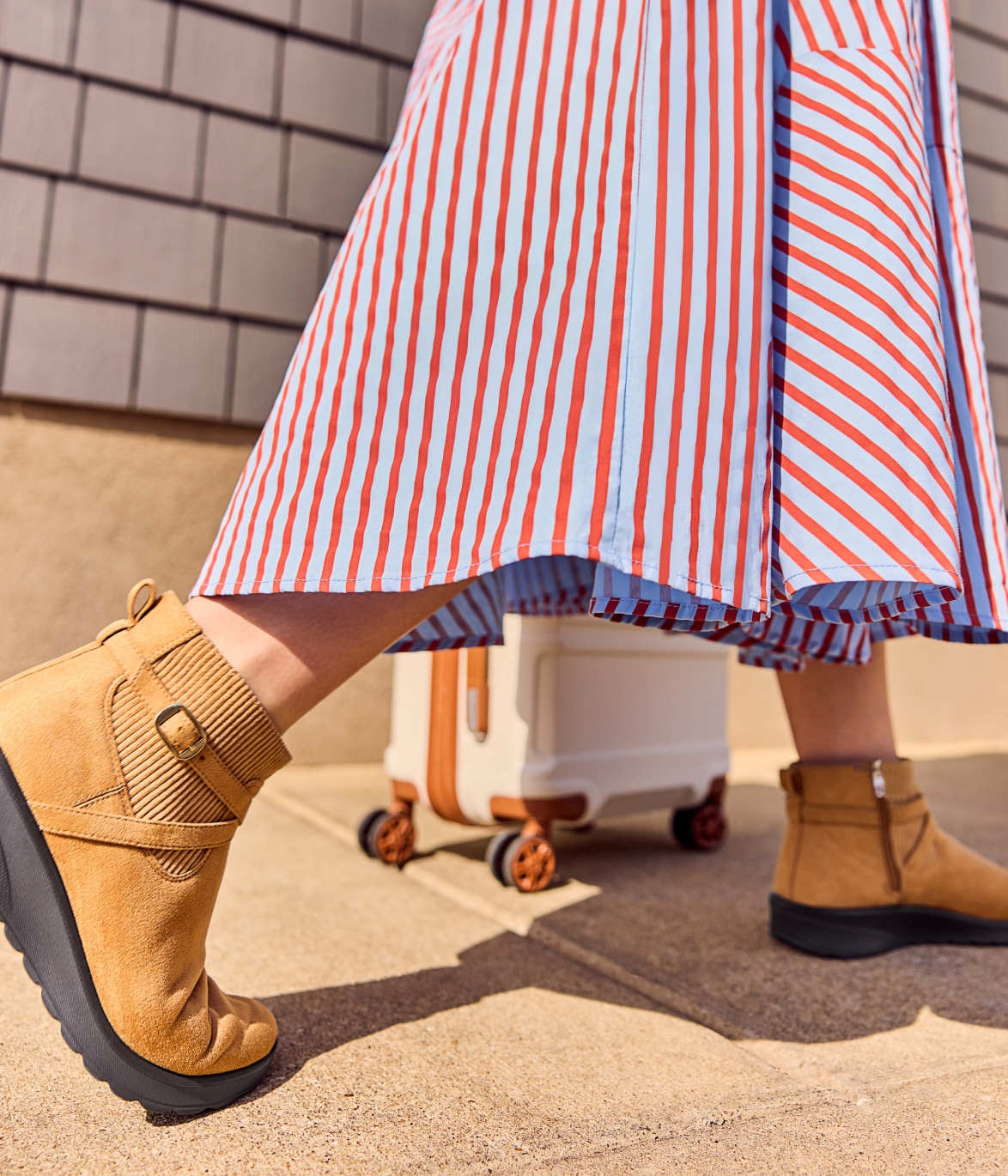 a model wearing a long striped skirt and taupe LifeStride travel style shoes while pulling a rolling suitcase 