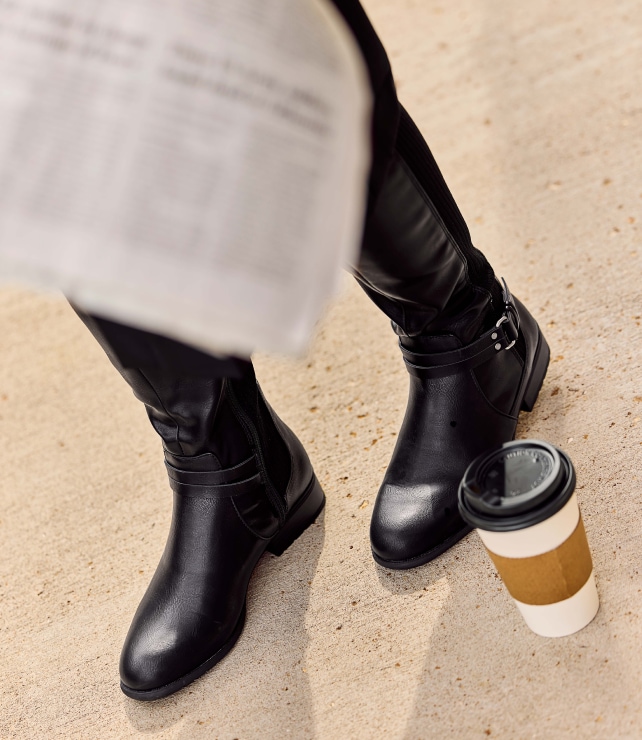a model wearing black LifeStride boots standing next to a coffee cup while a newspaper hangs in the shot