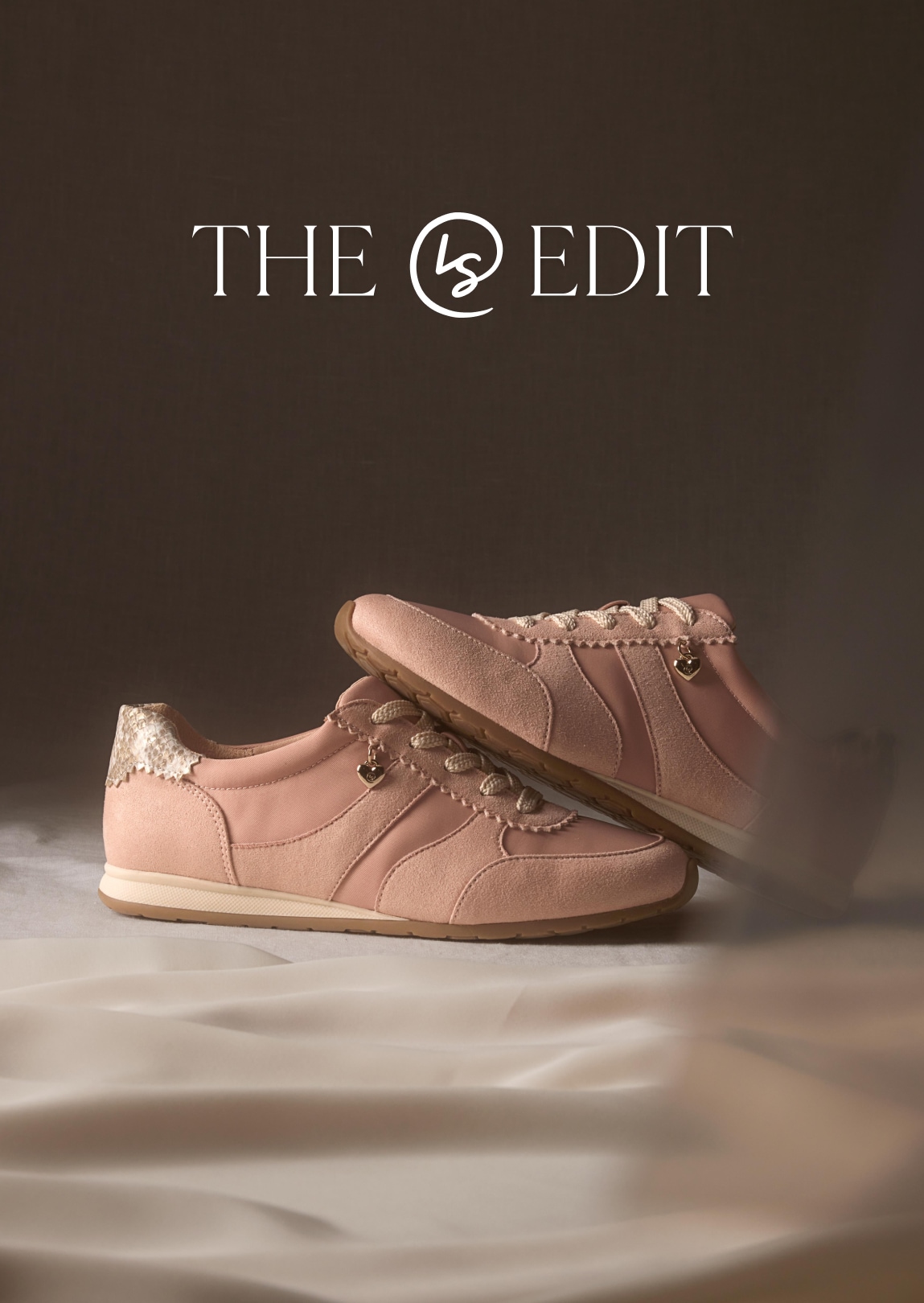 A pair of blush pink sneakers with suede panels and metallic heel detail, set on a brown backdrop with draped fabric and the text “THE LS EDIT” above.