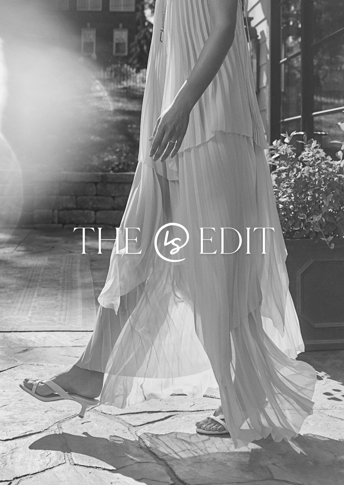 Black-and-white fashion photo of a woman wearing a flowy pleated dress and heeled sandals with “THE LS EDIT” text overlaid.