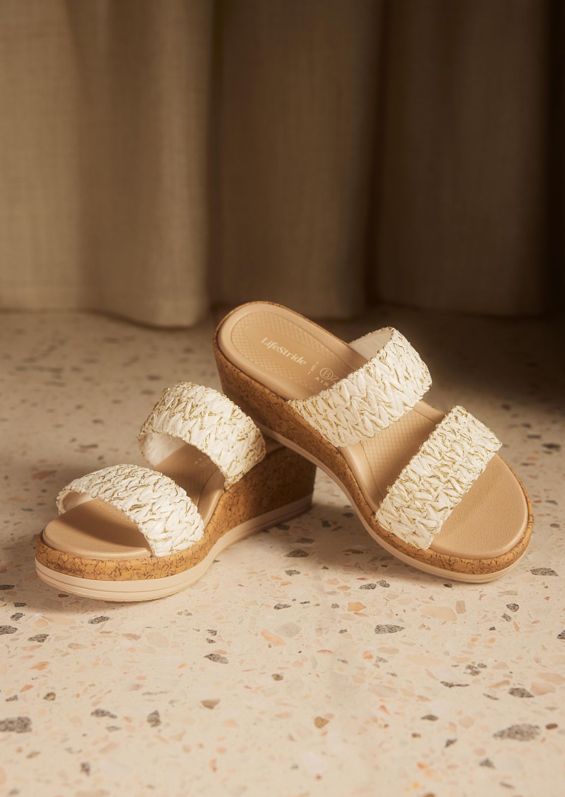 A pair of women's wedge sandals with a cork bottom and white and gold raffia straps displayed on a terrazzo floor.