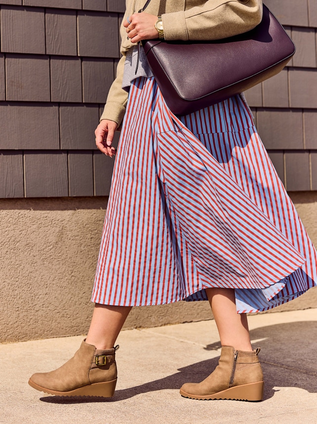 A model walking while carrying a large purple purse, wearing a taupe jacket and blue and red striped skirt and cozy LifeStride boots.