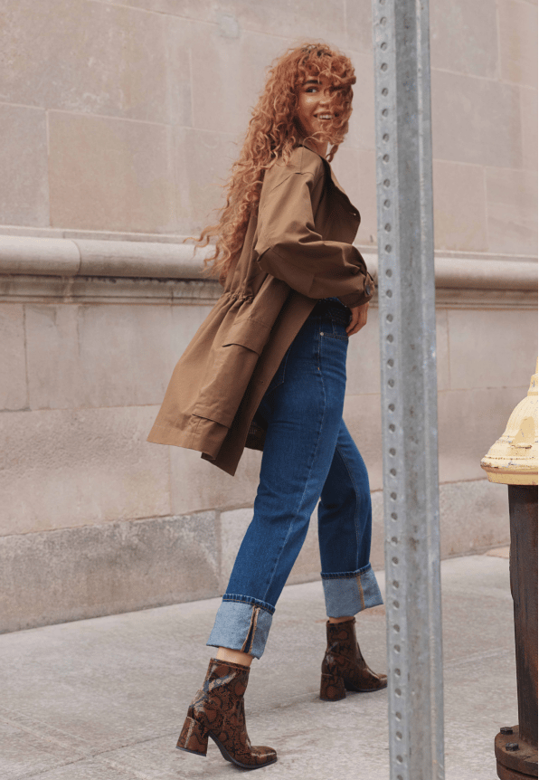 Woman walking down a sidewalk wearing a tan trench coat, jeans, and brown snakeskin boots.