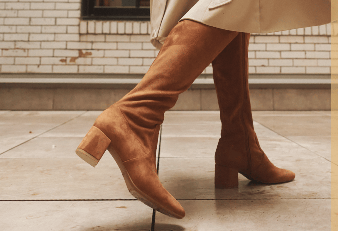 Woman wearing block heel knee high boots in tan microsuede fabric. 