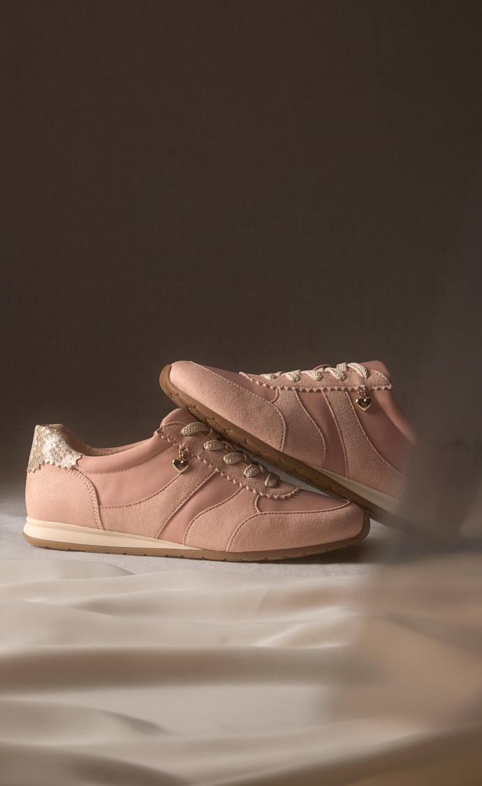 A pair of blush pink sneakers with suede panels and metallic heel detail, set on a brown backdrop with draped fabric.