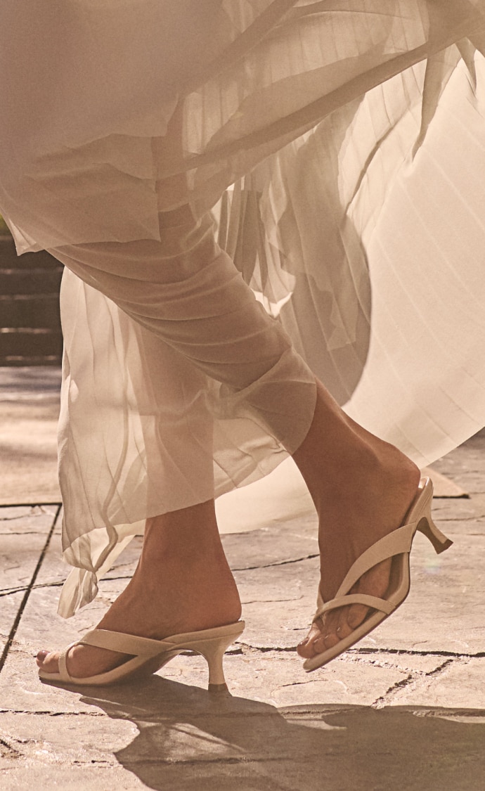 A closeup view of a woman wearing a flowy white dress with cream-colored heeled sandals. 