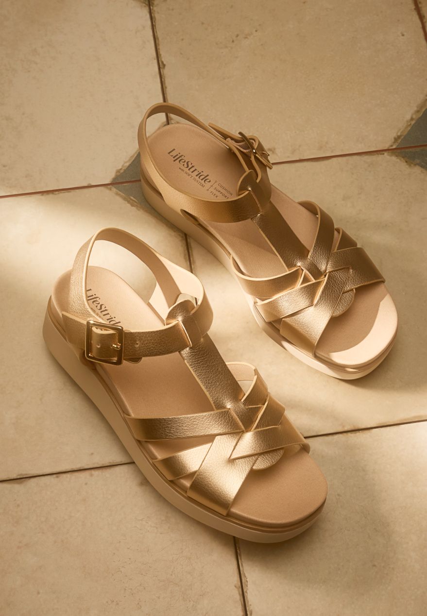 A pair of Lifestride's gold metallic Grandiose strappy wedge sandals on a beige and brown tile floor.