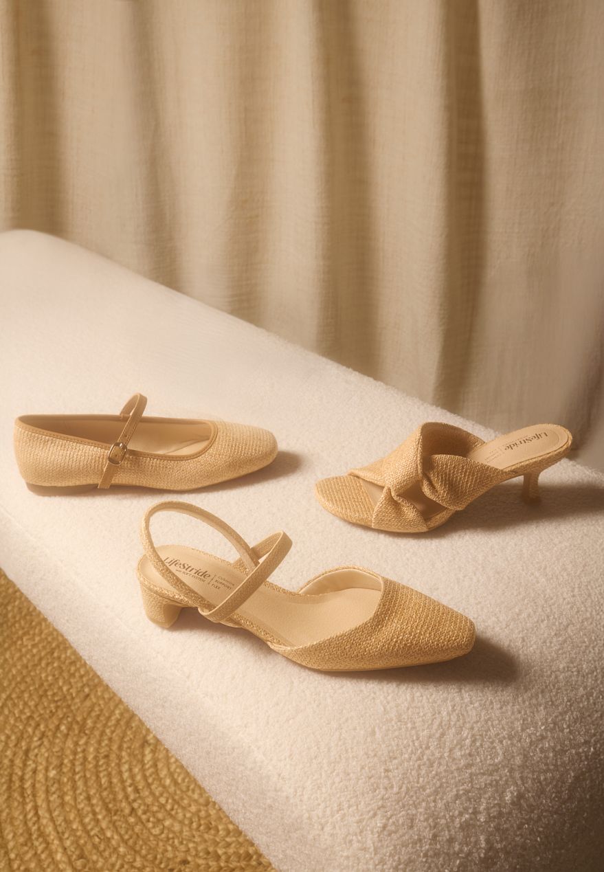 Three women's raffia shoes, including a Mary Jane flat and a heel, positioned on a white fabric bench with a tan fabric backdrop. 