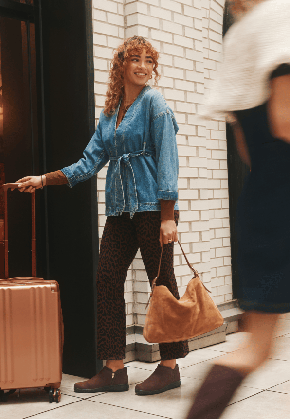 Woman standing next to luggage wearing a denim top, leopard print pants, and brown ankle boots. 